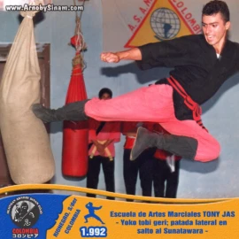 Arnoby Sinam realizando una yoko tobi geri (patada lateral en salto) sobre un sunatawara durante una sesión de entrenamiento en la Escuela de Artes Marciales Tony Jas. Arnoby Sinam ejecutando una yoko tobi geri, o patada lateral en salto, contra un sunatawara durante un entrenamiento en la Escuela de Artes Marciales Tony Jas en 1992.
