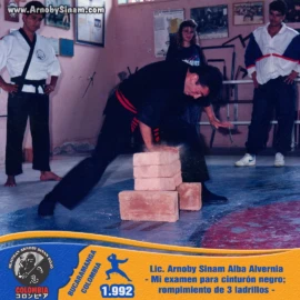Arnoby Sinam ejecutando la prueba de rompimiento de tres ladrillos durante su examen para cinturón negro, demostrando técnica, precisión y poder. Arnoby Sinam realiza el rompimiento de tres ladrillos durante su examen para cinturón negro en artes marciales en 1992.